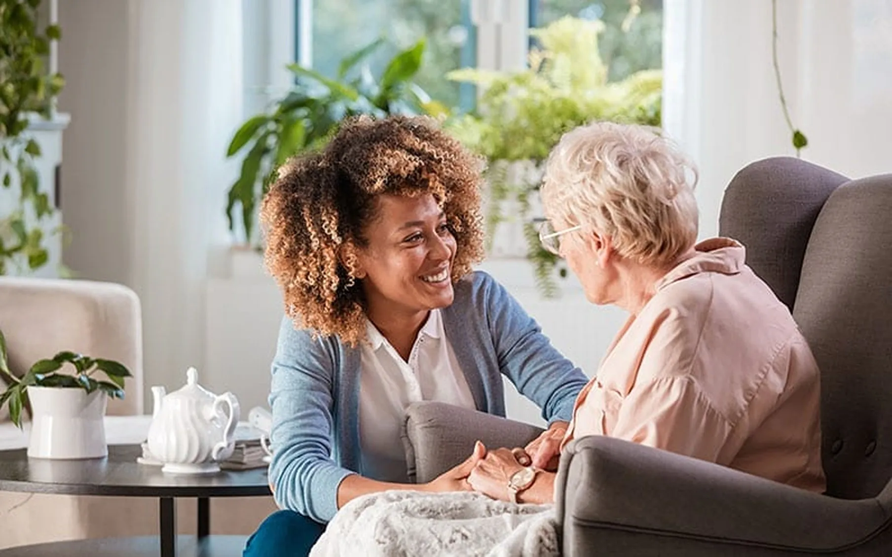 A carer smiling and holding the hands of an older woman as they talk in a bright living room