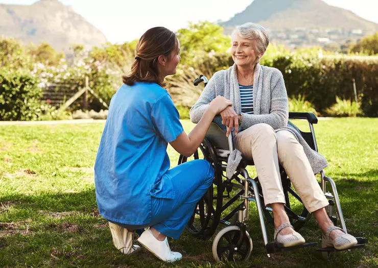 A carer crouching beside an older woman in a wheelchair, both smiling, on a lawn in the sunshine