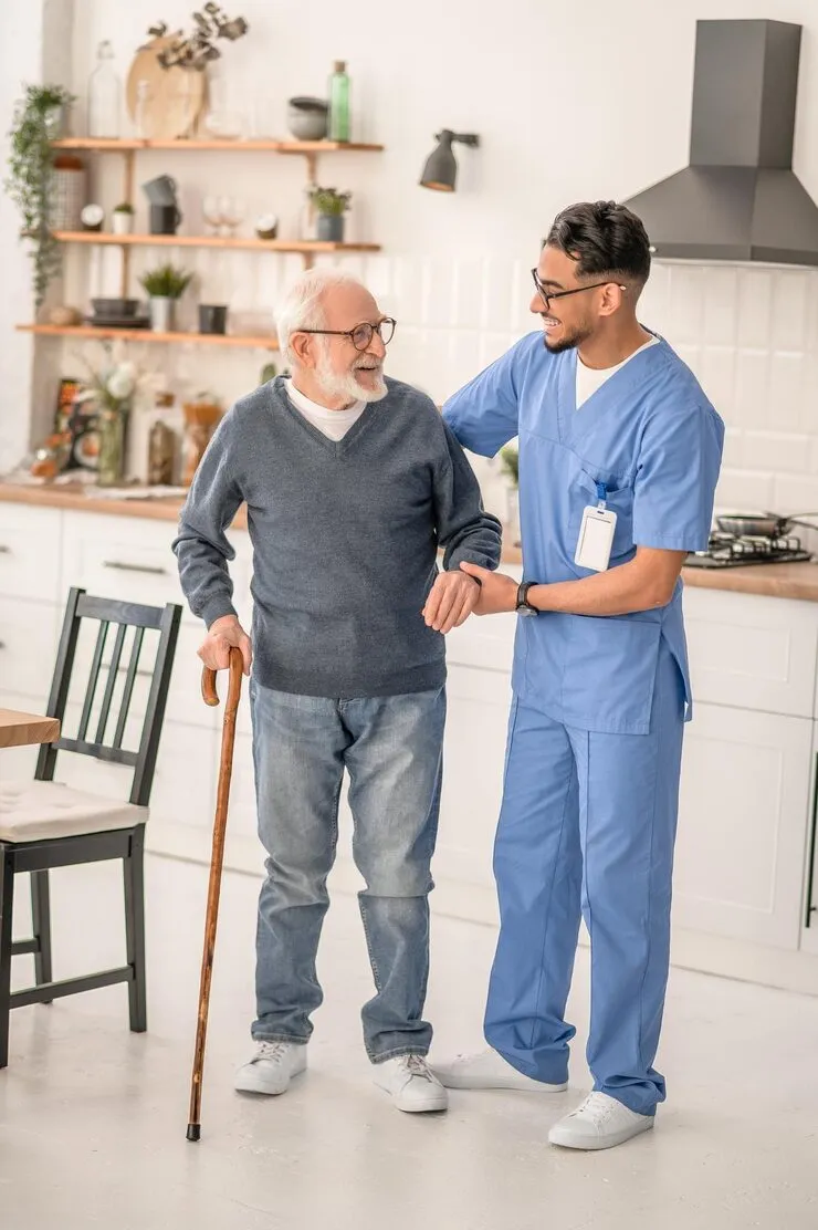 A carer in blue scrubs steadying an older man with a walking stick in a bright kitchen